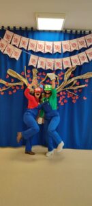 Na niebieskim tle z drzewem przyjaźni, dwie nauczycielki- organizatorki balu, w przebraniach postaci komputerowych Mario i Luigi.