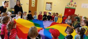 Grupa dzieci przedszkolnych przebrana w różne stroje karnawałowe wraz z nauczycielkami, bawiąca się przy kolorowej chuście animacyjnej.