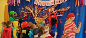 Grupa przebranych dzieci wraz z nauczycielami dekorują drzewo przyjaźni serduszkami z imionami dzieci z całego przedszkola.